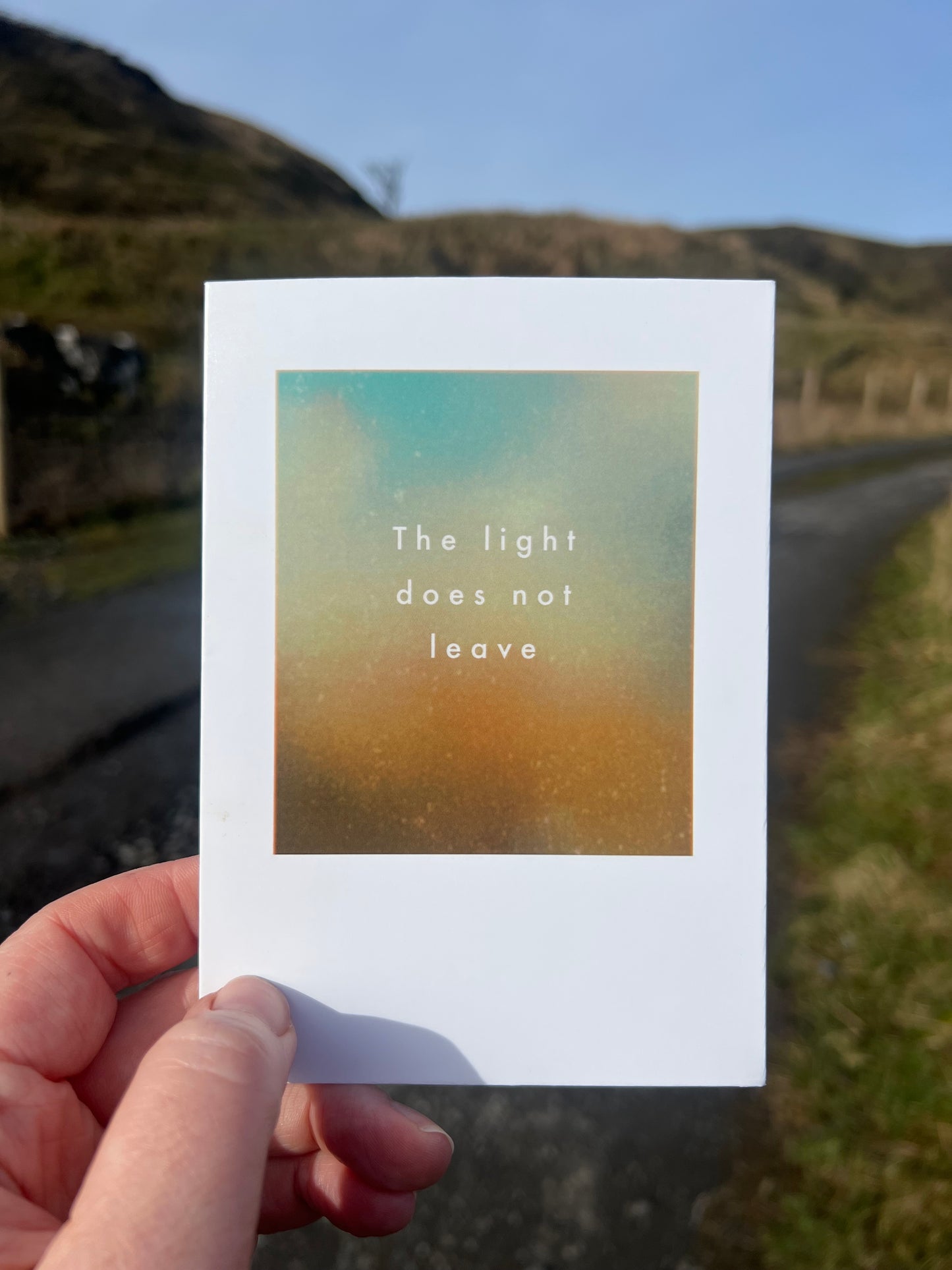 Hand holding a card with 'The light does not leave' text against a blurred natural background
