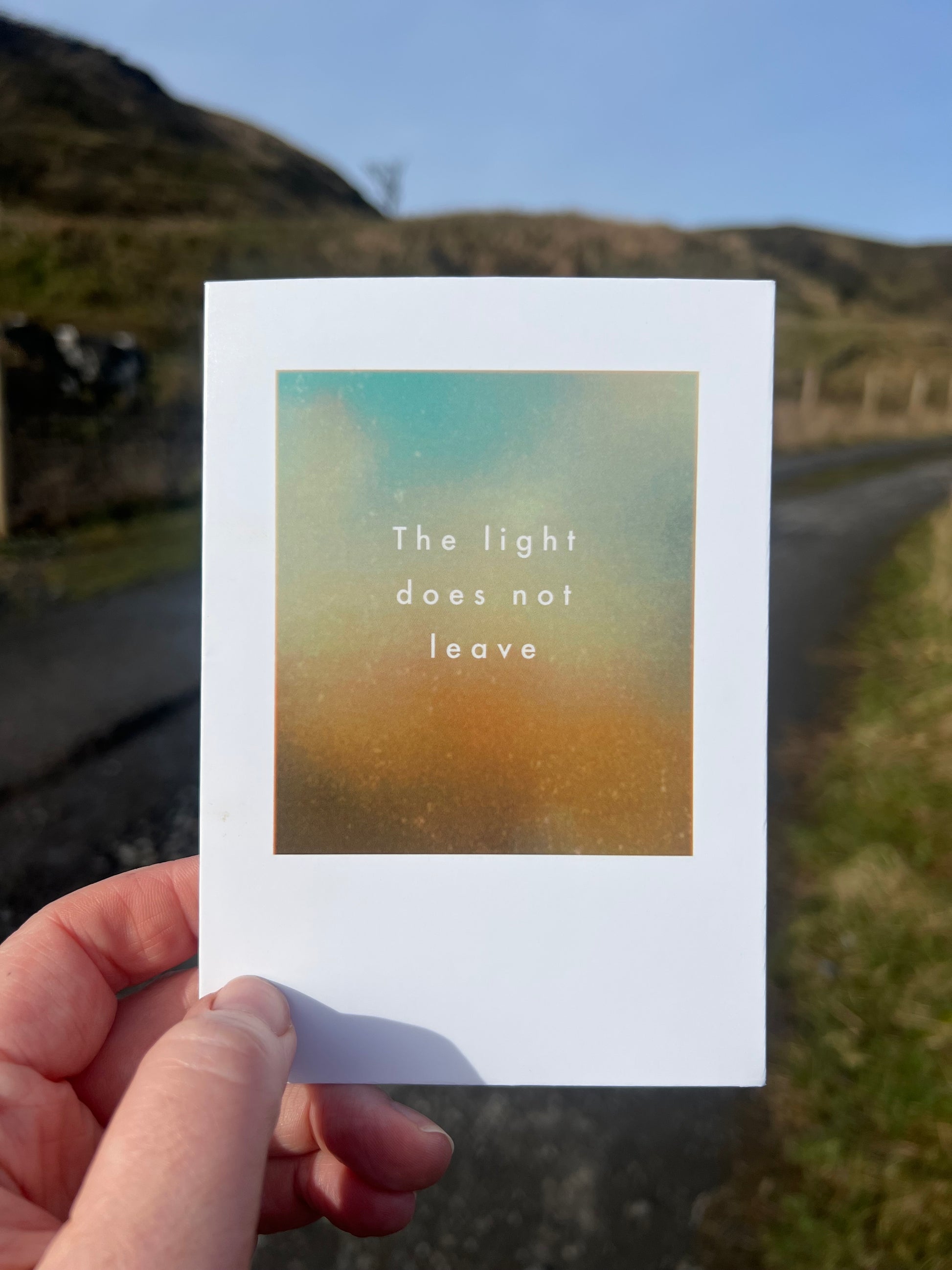 Hand holding a card with 'The light does not leave' text against a blurred natural background