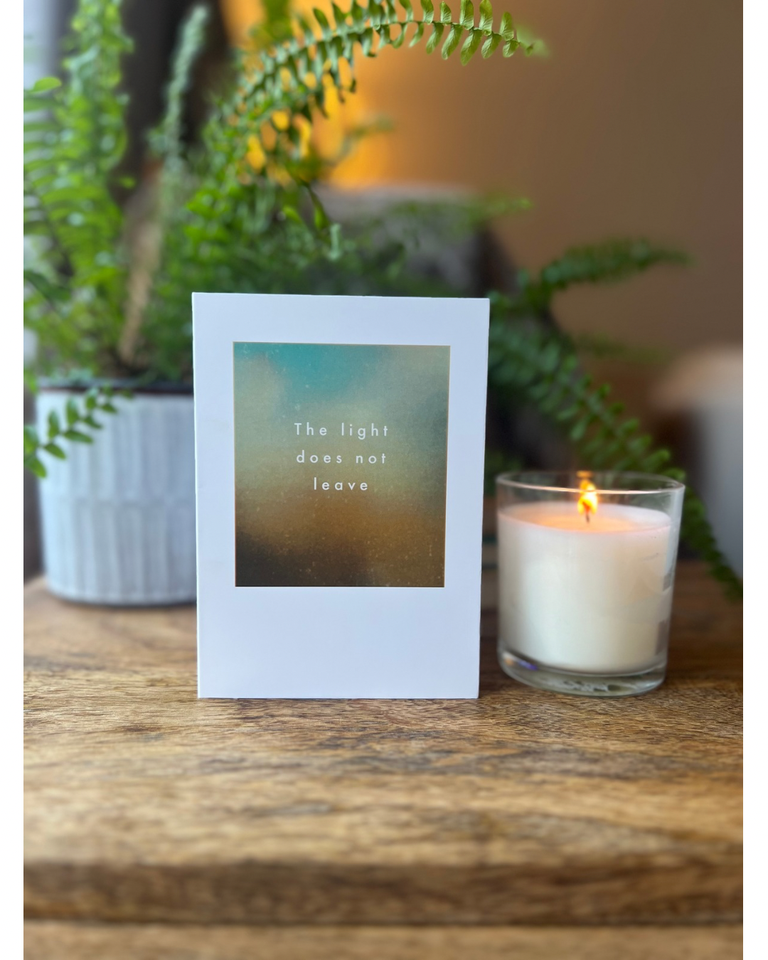 Candle with a card displaying 'The light does not leave' on a wooden surface with a plant in the background.