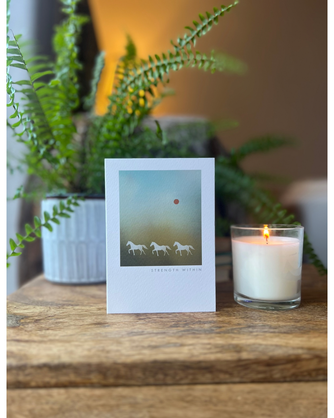 Greeting card with horses on a wooden surface next to a lit candle and plant.